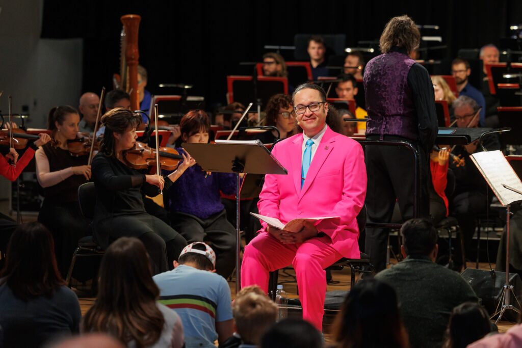 A man in a vibrant pink suit sitting in front of a group of violin players and a harpist talks to an audience of children and parents